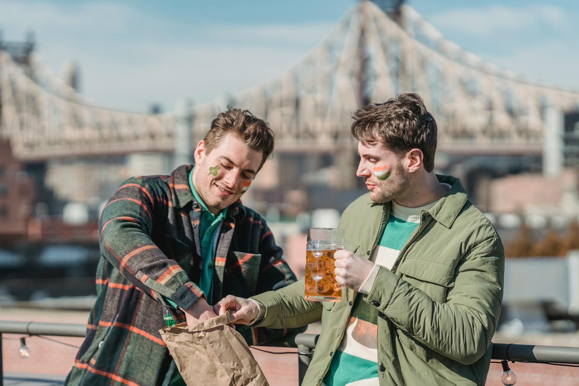 Male friends with shamrock and flag painted on faces standing on seafront with glass of beer and snacks on street