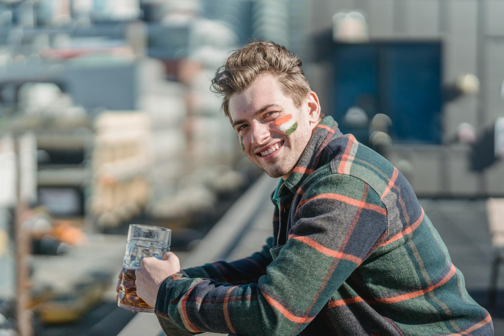 Smiling young male in colorful coat with painted Irish flag on cheek standing on rooftop with glass of beer on blurred background