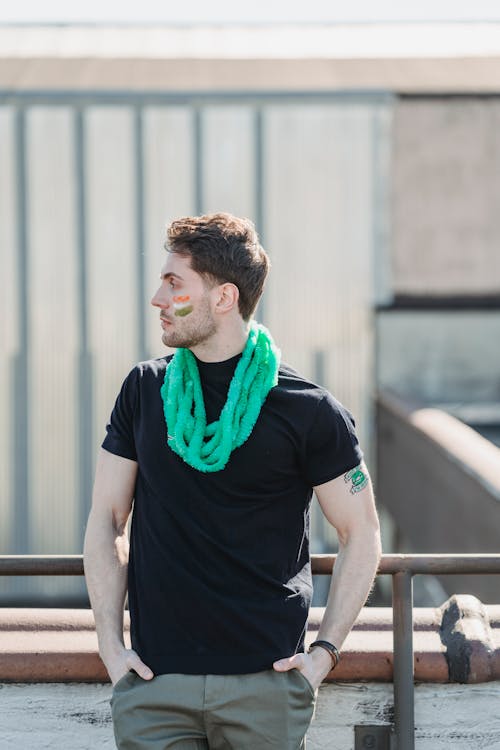 Confident young male in casual outfit and festive necklace with painted flag of Ireland on face standing on balcony with hands in pockets and looking away on St Patricks Day