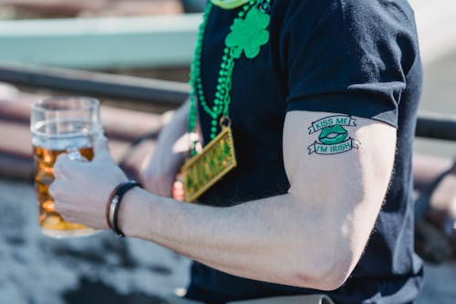 Side view of crop anonymous male with creative Kiss Me Im Irish tattoo on arm in casual clothes and shamrock necklace drinking mug of cold beer during St Patricks Festival