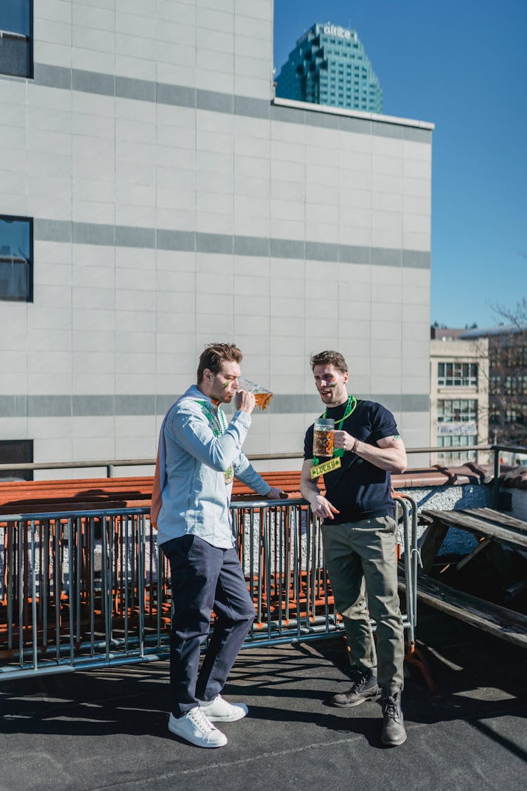 Young Men Drinking Mugs Of Beer While Celebrating Irish Holiday On Terrace