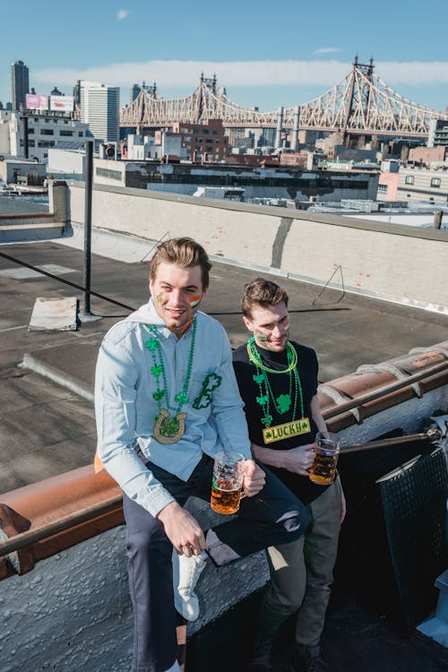 From above of positive young male friends in casual outfits and festive shamrock necklaces smiling and drinking beer while chilling on building roof during celebration of St Patricks Day