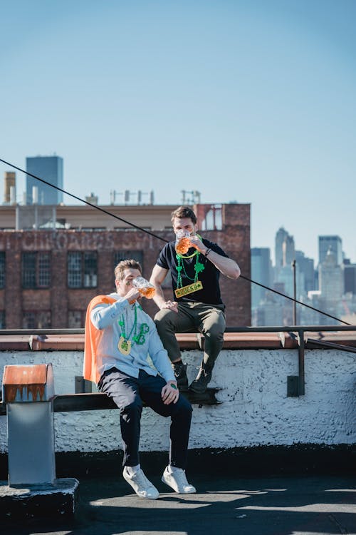 Full body of young male friends with festive necklaces and flag on shoulders boozing beer on building rooftop while celebrating Feast of St Patrick on sunny day