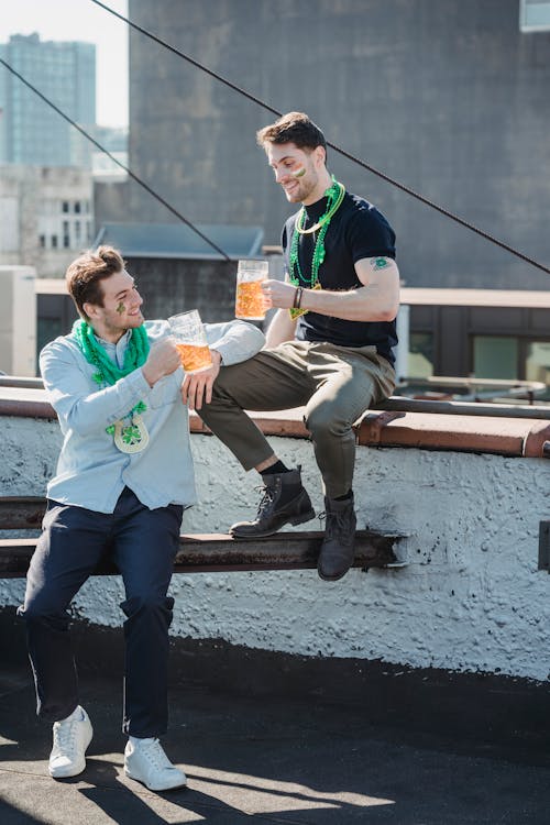 Full body of cheerful young male friends with painted faces and festive accessories clinking glasses of beer while celebrating Feast of Saint Patrick on rooftop of multistory building