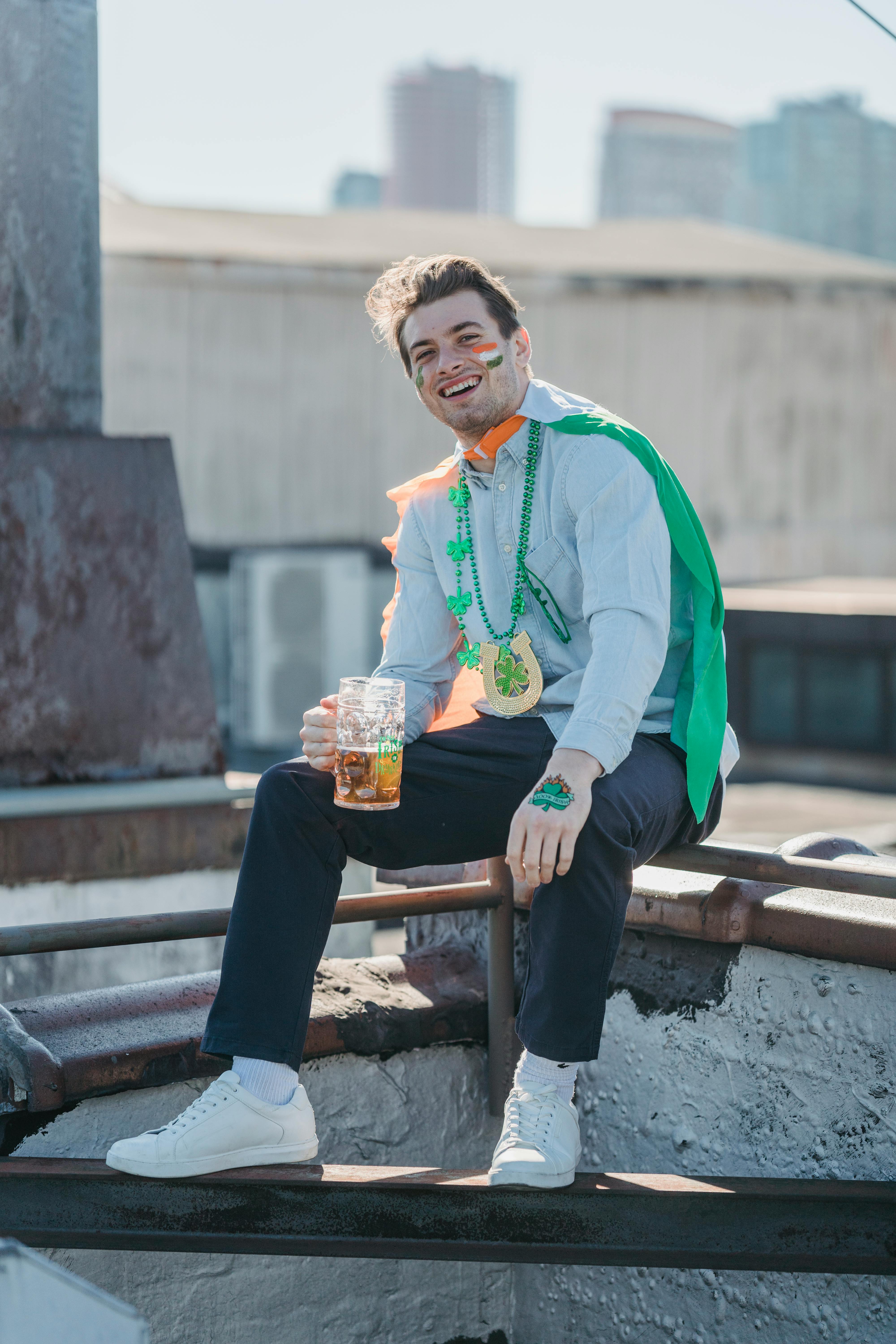 Full body of cheerful young male in stylish clothes and flag of Ireland on shoulder smiling and looking at camera while drinking beer on rooftop on St Patricks Day