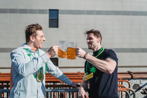Free Excited young male best friends with festive accessories clinking mugs of beer while celebrating St Patricks Day on street on sunny day Stock Photo