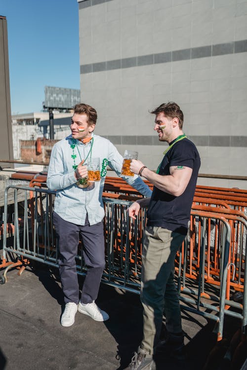 Happy young male friends in casual clothes with painted faces smiling an looking away while drinking beer on terrace during St Patricks Day party