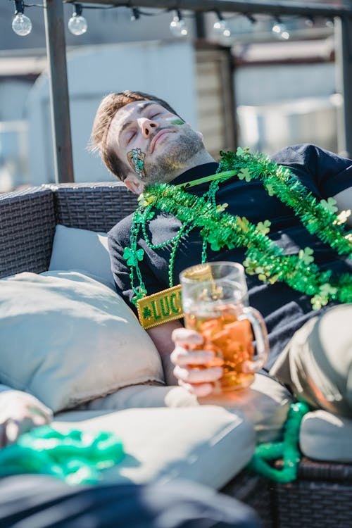 Free Drunk young unshaven guy with painted face and green festive tinsels lying on sofa with closed eyes and beer mug in hand after after outdoor party on St Patricks Day Stock Photo