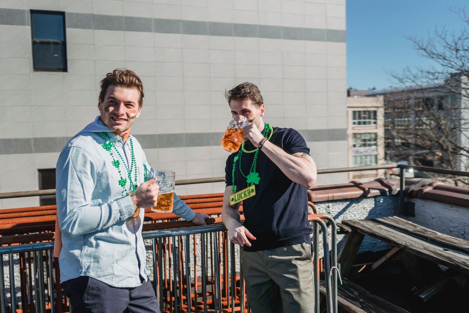 Happy young male millennials in casual clothes and clover necklaces drinking glasses of beer while celebrating Feast of Saint Patrick on sunny day on terrace Happy young male millennials in casual clothes and clover necklaces drinking glasses of beer while celebrating Feast of Saint Patrick on sunny day on terrace