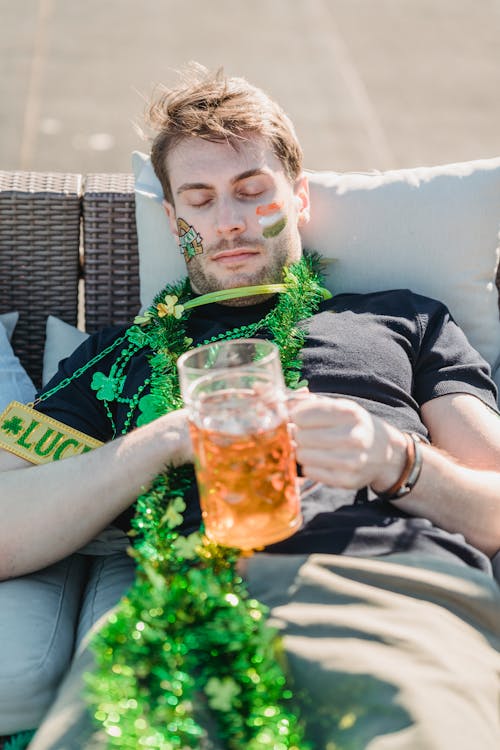 Free High angle of drunk young man with painted face and festive accessories lying on couch with mug of beer in hands and sleeping after hangout on St Patricks Day Stock Photo