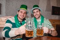 Cheerful young friends celebrating Saint Patrick day wearing national green hat and scarf raising mug of beer in pub