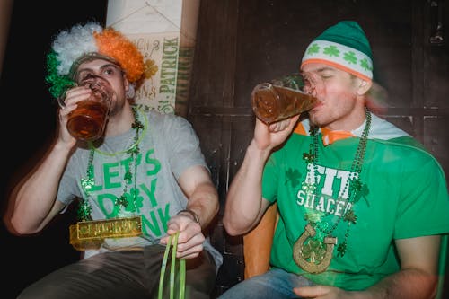 Delighted young friends wearing Irish national symbols emerald green hat and party wig in flag colors and necklace and neon green glow sticks drinking beer while enjoying Saint Patrick Day celebration