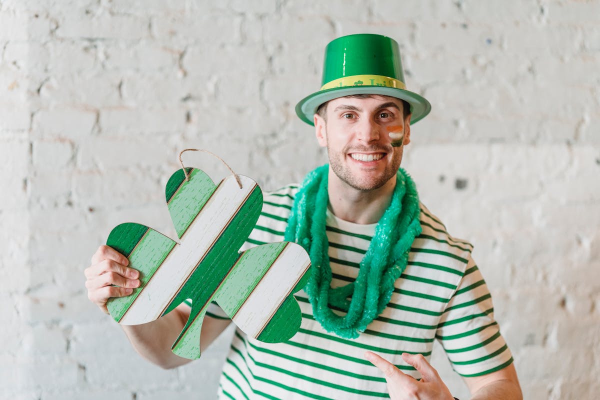 Amusing young man with colored in Irish flag cheeks wearing green hat and green sparkle scarf with pasteboard gift in form of Irish symbol shamrock celebrating St Patricks Day