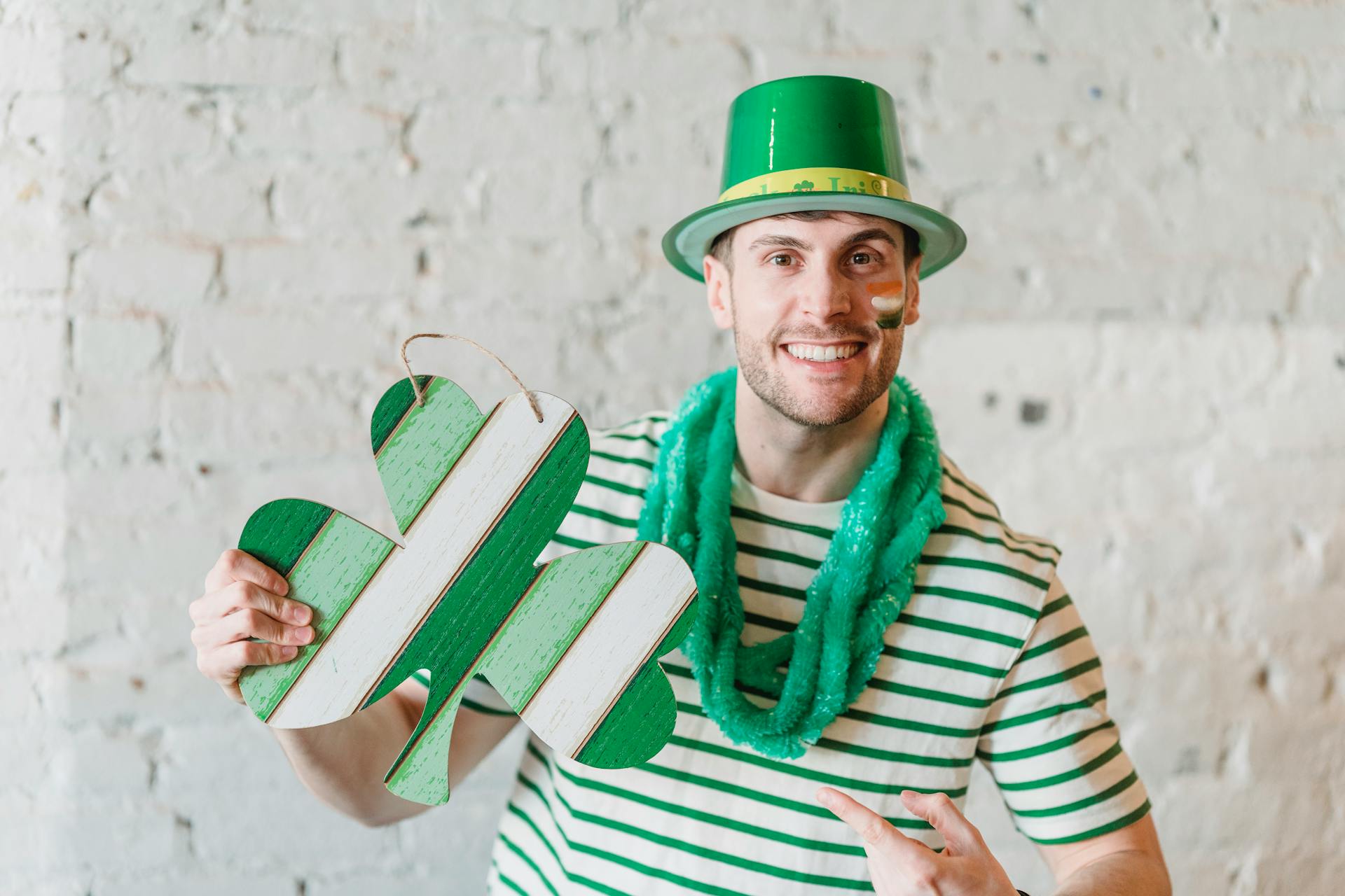 Grappige jonge man met gekleurde in Ierse vlag wangen draagt een groene hoed en groene glinsterende sjaal met karton geschenk in de vorm van een Ierse symbool klaver vieren St Patrick's Day