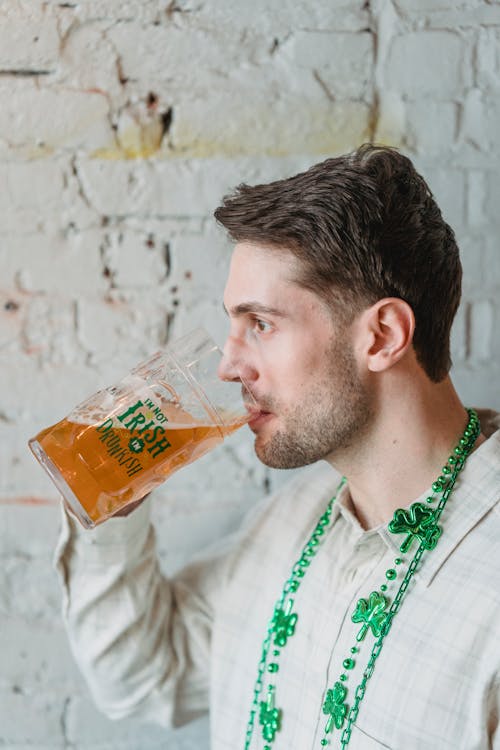 Side view of concentrated unshaven young guy wearing necklace decorated with shamrock and green sparkle beads drinking large mug of beer in honor of St Patricks Day