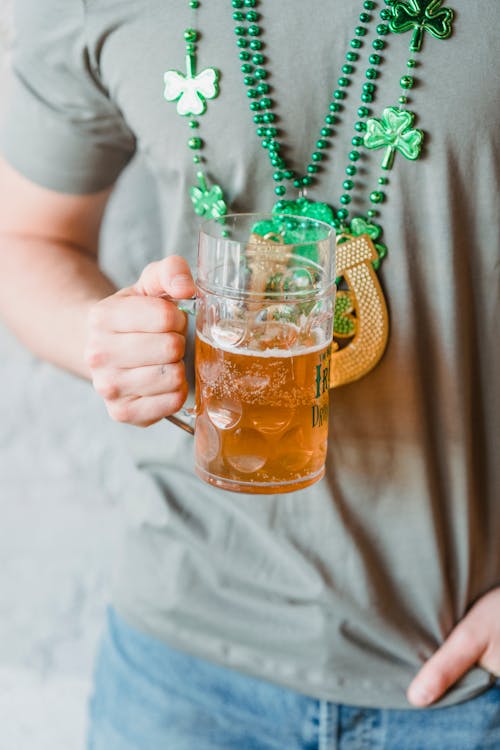 Crop unrecognizable person holding mug of beer and wearing gray casual t shirt and blue jeans and necklace with elements of green shamrock and wooden symbol of horseshoe