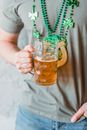 Crop unrecognizable person holding mug of beer and wearing gray casual t shirt and blue jeans and necklace with elements of green shamrock and wooden symbol of horseshoe