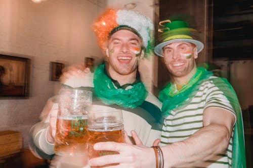 Cheerful men covered flag of Ireland and wearing green necklace and green national hat gathering in pub while drinking beer and celebrating Saint Patricks Day