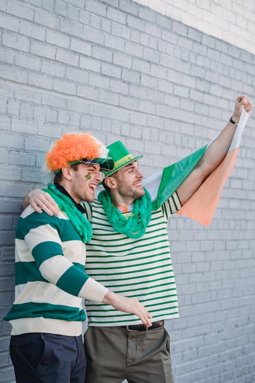 Side view of male friends wearing clown party wig and green stripe t shirt and green sparkle hat covered with irish flag greeting and celebrating Saint Patrick Day