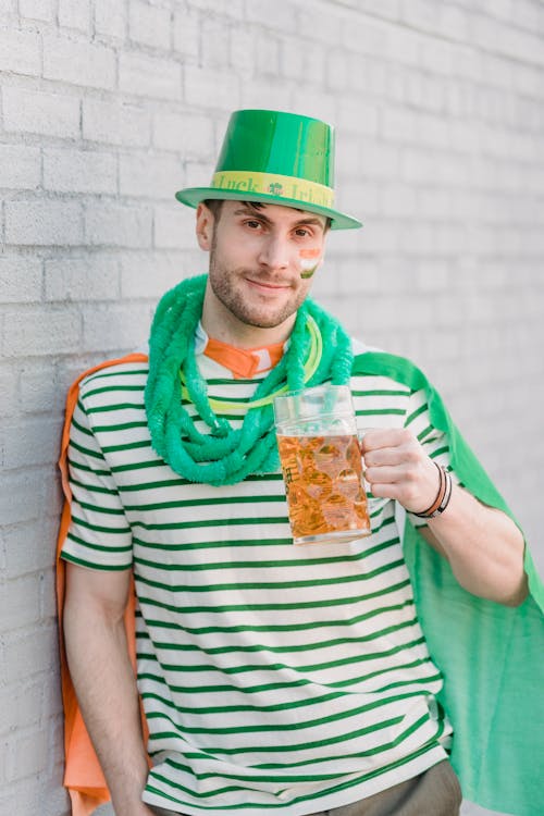 Smiling bearded young male in green necklace and green hat with flag of Ireland on shoulders celebrating Saint Patricks Day