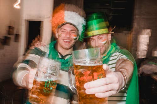 Laughing friends enjoying holiday with beer in pub wearing clown party wig of Irish national flag colors green hat and covered with cloth of Irish national flag colors