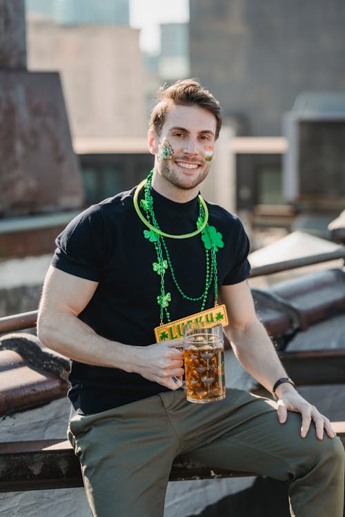 Cheerful young bearded male with painted face in casual clothes and festive shamrock necklace smiling and looking at camera while drinking beer on building roof during St Patricks Day celebration