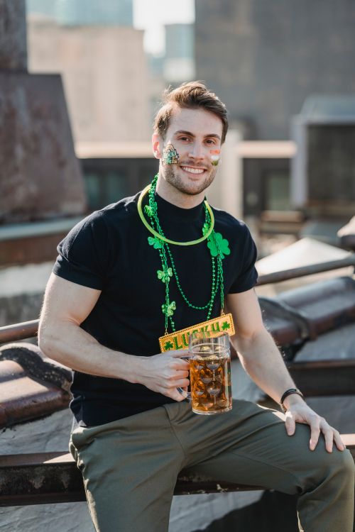 Free Cheerful young bearded male with painted face in casual clothes and festive shamrock necklace smiling and looking at camera while drinking beer on building roof during St Patricks Day celebration Stock Photo