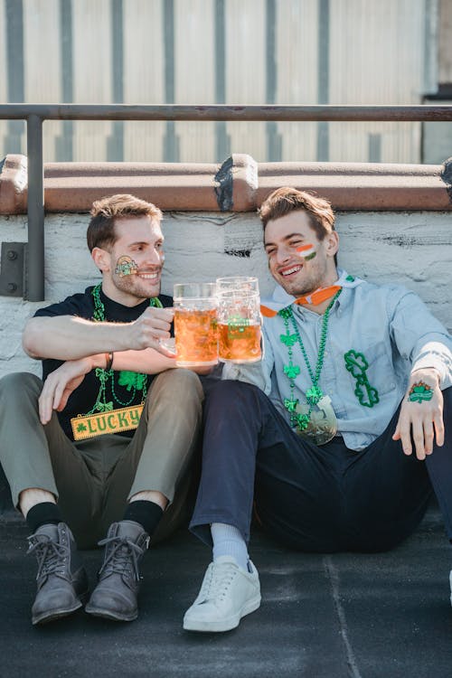 Cheerful young male best friends with painted faces clinking glasses of beer and smiling while sitting on building rooftop and celebrating Feast of Saint Patrick