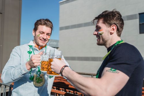 Free Delighted young unshaven guys in casual clothes and festive accessories toasting with beer glasses and smiling while celebrating St Patricks Day together on terrace Stock Photo