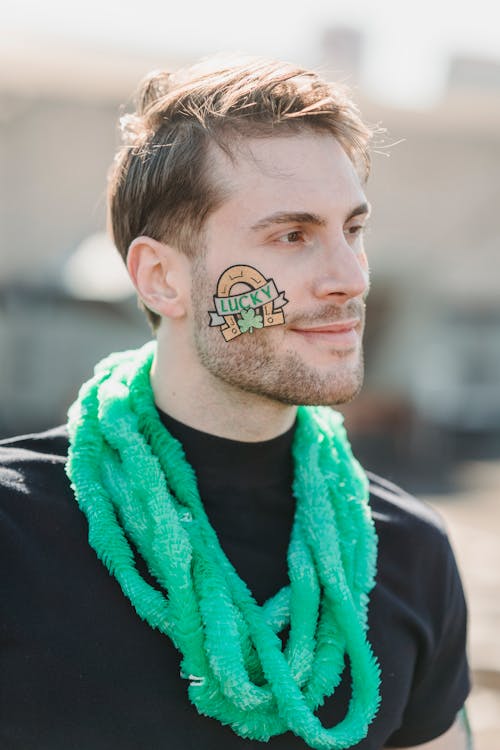 Free Smiling young unshaven guy with lucky sticker on cheek and festive necklace looking away during celebration of St Patricks Day Stock Photo