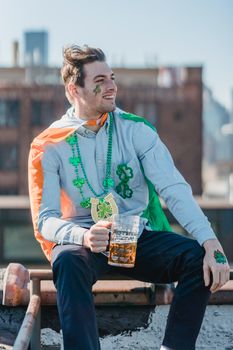Joyful young man in casual clothes with Irish flag on shoulders smiling while drinking glass of beer on building rooftop on St Patricks Day