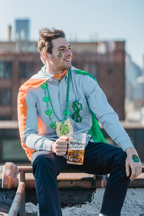 Free Joyful young man in casual clothes with Irish flag on shoulders smiling while drinking glass of beer on building rooftop on St Patricks Day Stock Photo