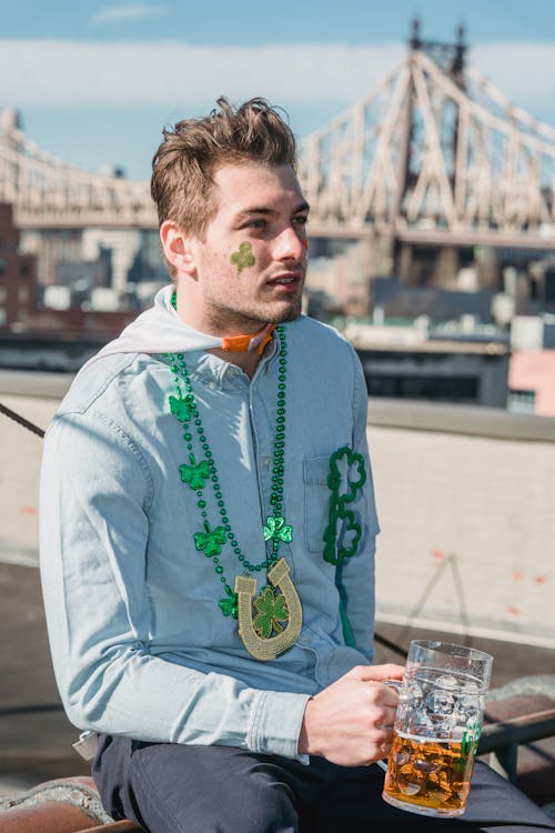 Free Confident young unshaven male millennial in casual clothes with festive accessories drinking glass of beer and looking away pensively during celebration of Feast of Saint Patrick Stock Photo