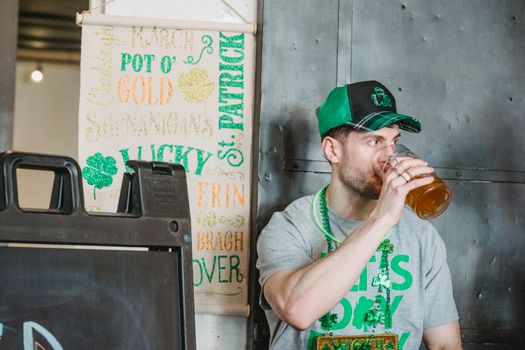 Young guy in festive outfit drinking glass of beer and looking away during celebration of St Patricks Day in pub