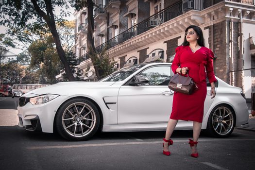 Fashionable woman in red dress holding handbag next to a luxury white car on city street.