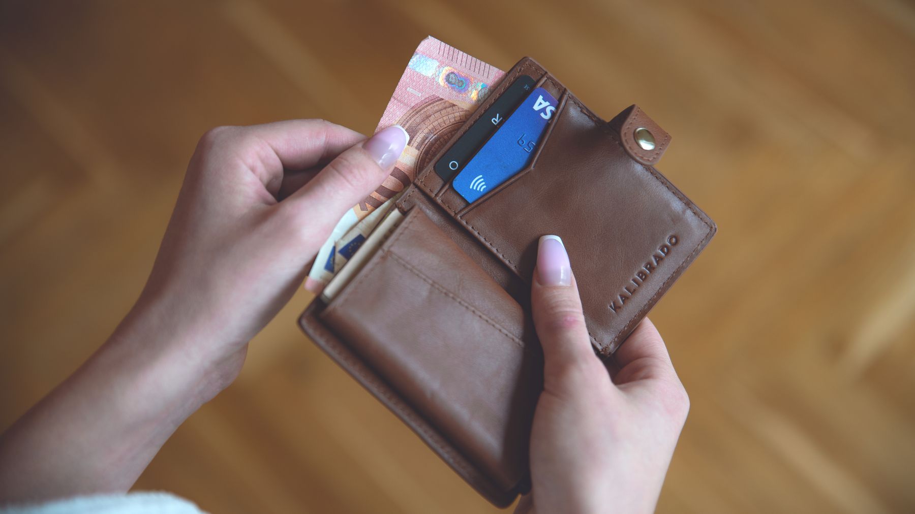 Hands holding a leather wallet with cash and a credit card on a wooden surface.