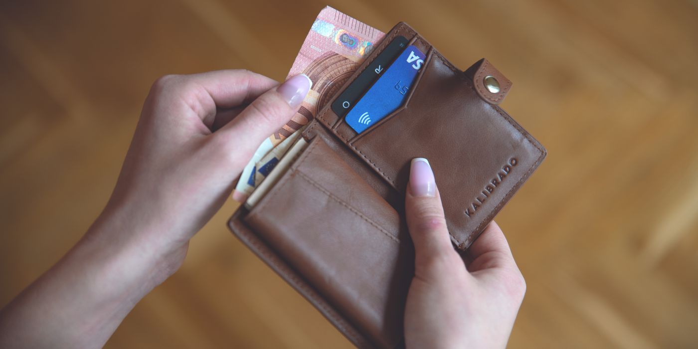 Hands holding a leather wallet with cash and a credit card on a wooden surface.