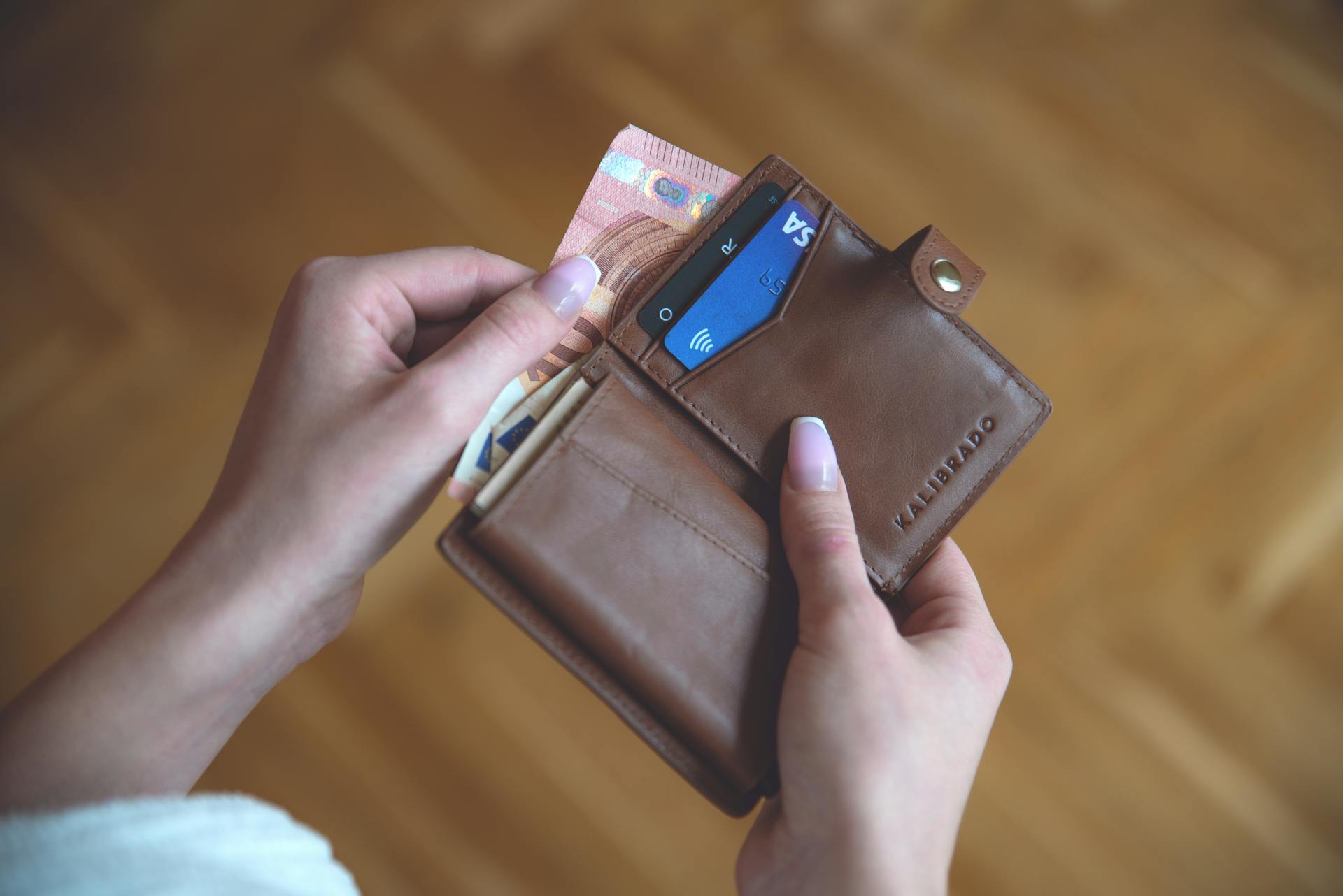 Hands holding a leather wallet with cash and a credit card on a wooden surface.