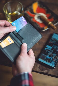 Hands holding a sleek black wallet containing credit cards next to a smartphone on a wooden table.