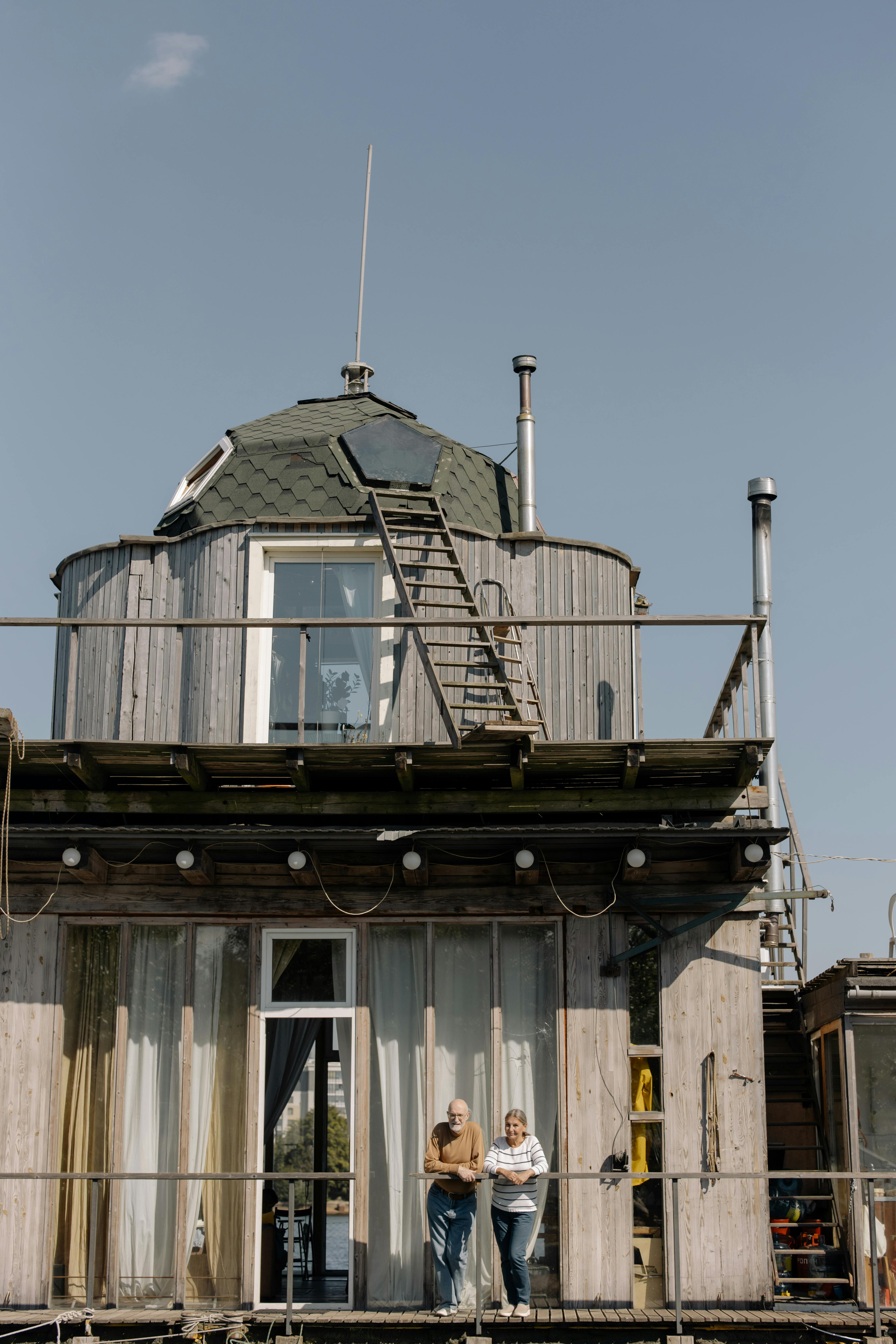 Seniors enjoying a sunny day on the deck of a unique wooden houseboat with a clear blue sky.