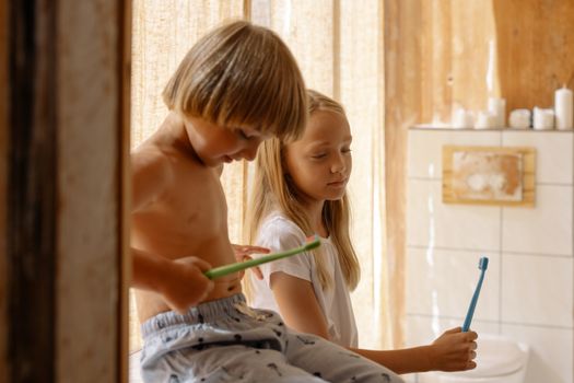 Two children brush their teeth in a warm bathroom, holding green and blue toothbrushes beside a sink.