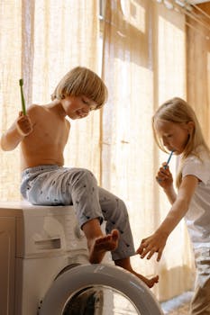 Two kids brushing teeth indoors on washing machine in natural light.