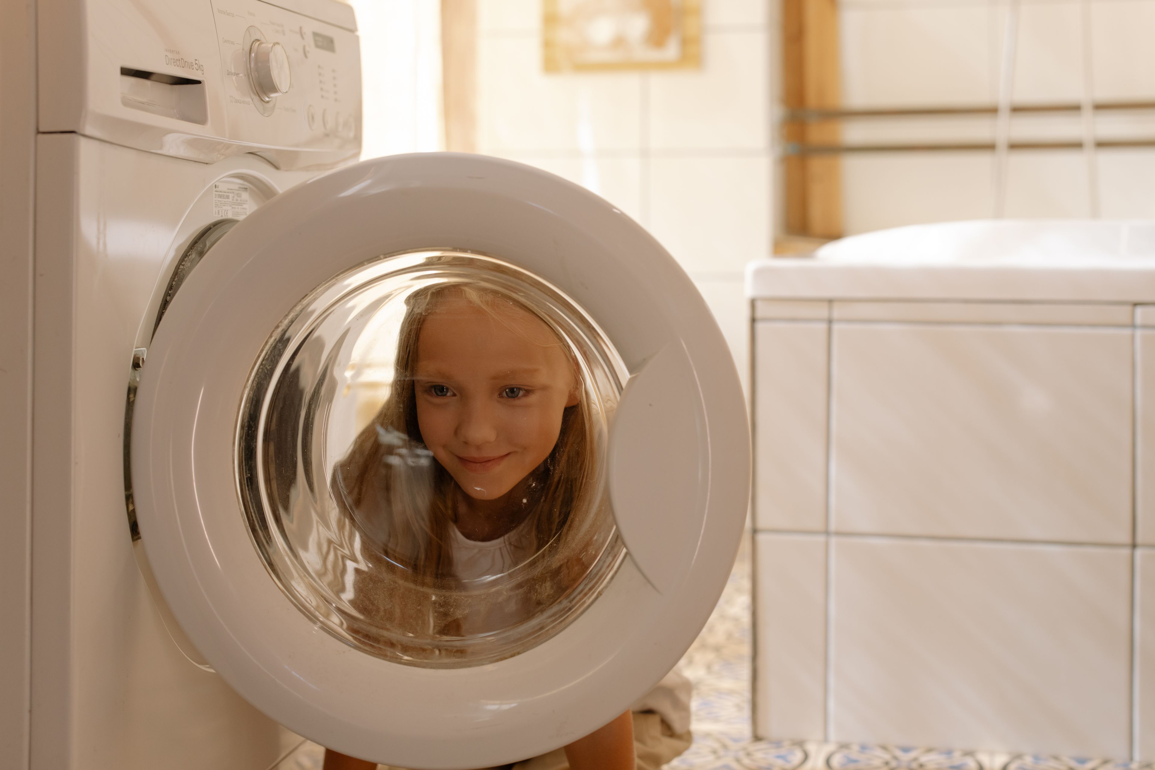 A young girl looks curiously into the washing machine door in a bright, modern bathroom.