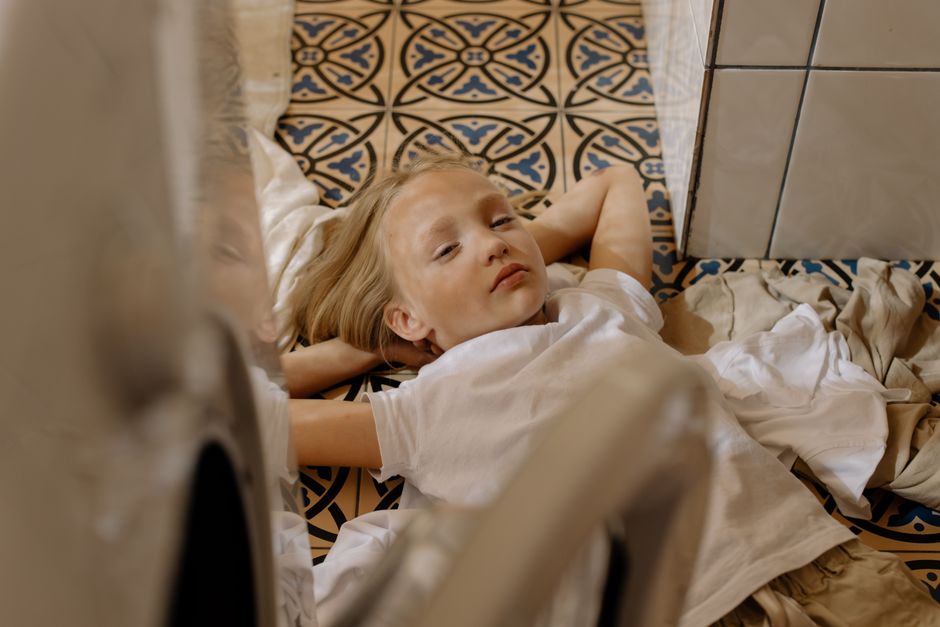 Photo by cottonbro studio on Pexels A blonde child lies comfortably on a patterned floor, surrounded by laundry indoors. - Photo by cottonbro studio on Pexels