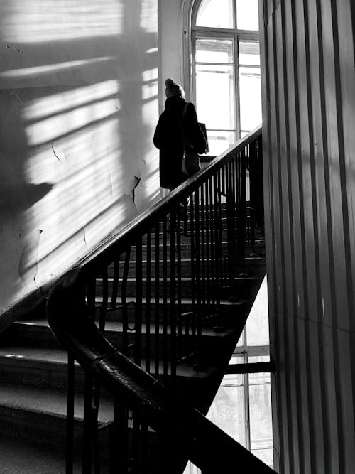 Black and white back view of unrecognizable person in outerwear walking upstairs in residential building near window with bright sunlight