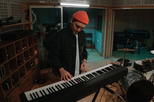 A musician wearing a beanie plays a keyboard in a cozy recording studio setting.