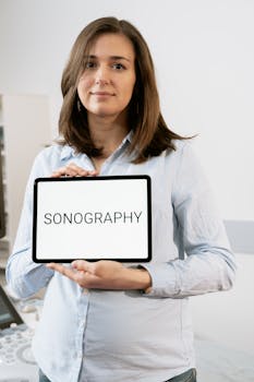 A pregnant woman holding a tablet with 'SONOGRAPHY' text inside a medical clinic.