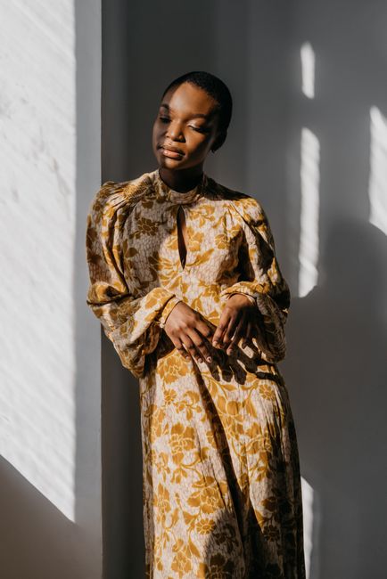A stylish woman in a floral dress posing gracefully in natural light indoors.