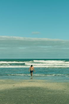 A shirtless man walks along a sunny beach with waves in the background, creating a serene seaside scene.