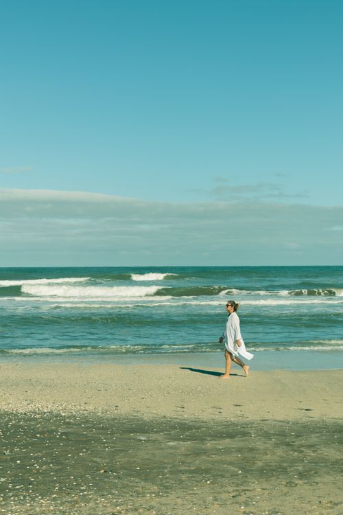 Free A woman in white walks barefoot along a serene beach under a clear blue sky. Stock Photo
